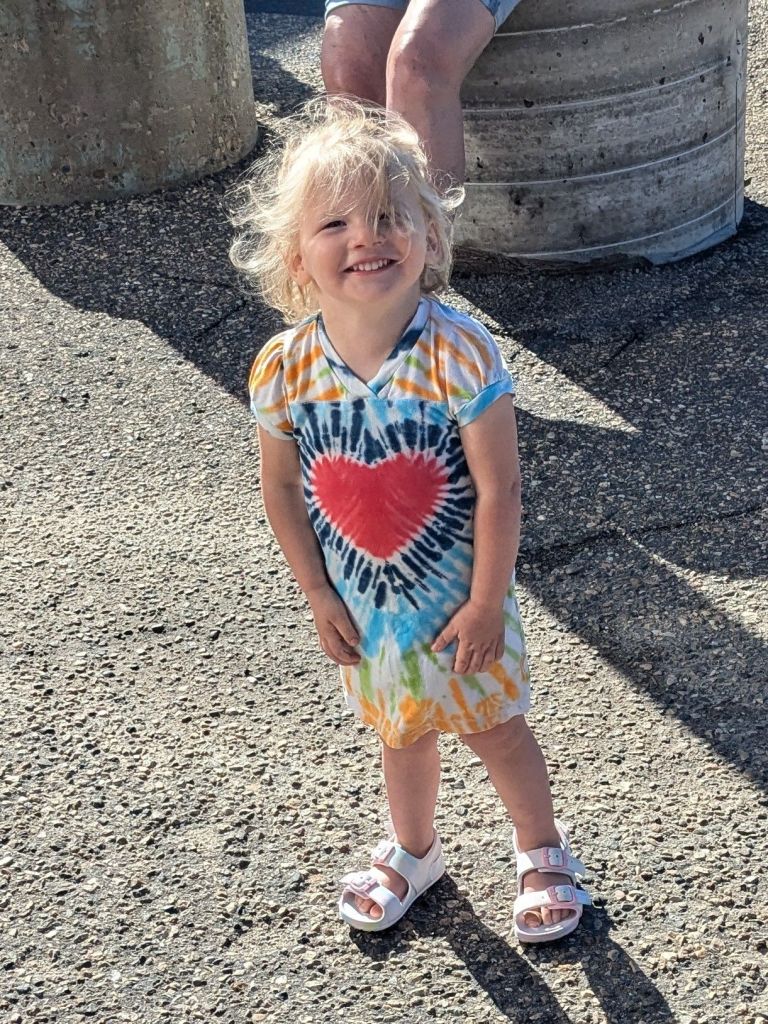 A joyful young child with messy hair smiles brightly while wearing a colorful tie-dye dress with a heart design, standing on a sunny surface.